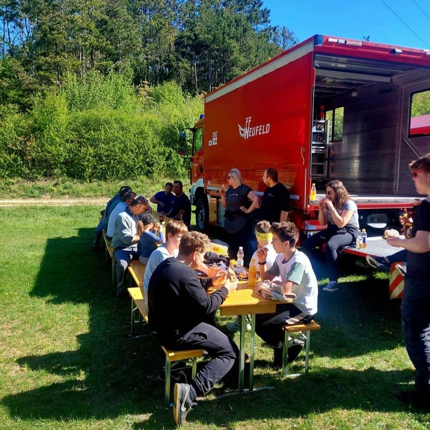 Eine Gruppe von Menschen isst an Picknicktischen im Freien in der Nähe eines roten Food Trucks mit dem Wort Neufeld. Sie sitzen auf einer Wiese mit Bäumen im Hintergrund.