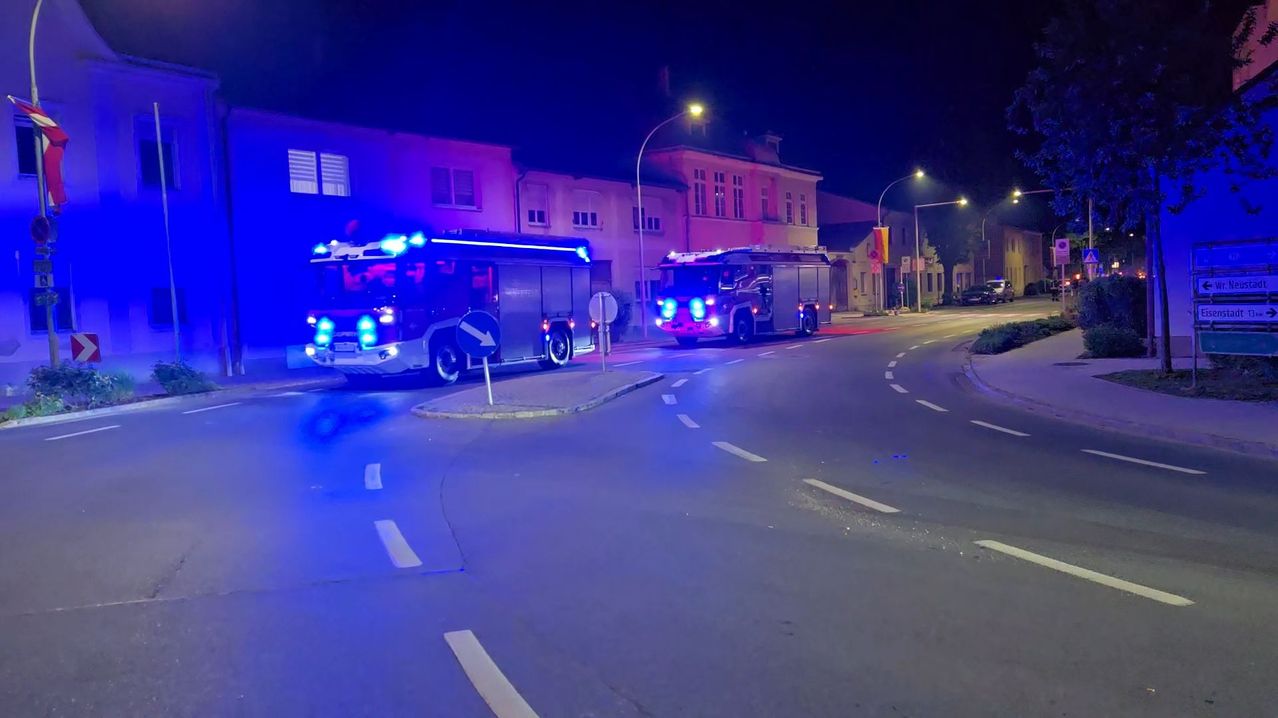 Zwei Feuerwehrwagen sind nachts am Straßenrand geparkt. Sie haben ihre blauen Lichter blinken. Ein Gebäude mit vielen Fenstern ist dahinter.