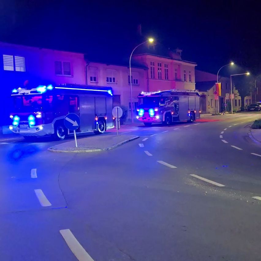 Zwei Feuerwehrwagen sind nachts am Straßenrand geparkt. Sie haben ihre blauen Lichter blinken. Ein Gebäude mit vielen Fenstern ist dahinter.