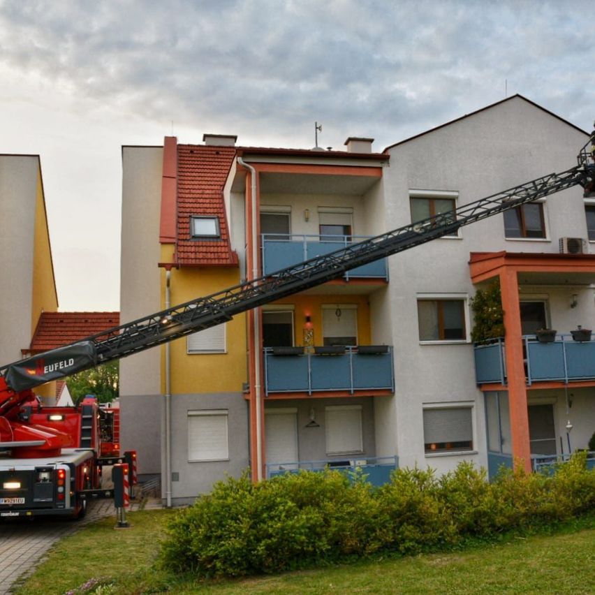 Ein Feuerwehrauto mit einer ausgefahrenen Leiter, die auf ein Mehrfamilienhaus mit Balkonen zeigt, unter einem bewölkten Himmel.
