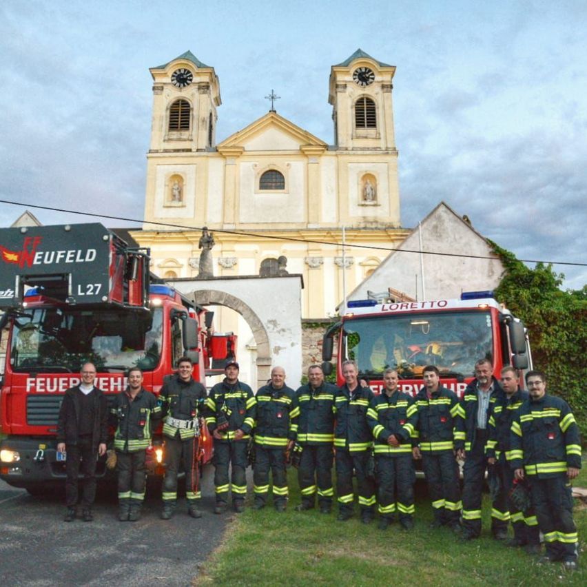Eine Gruppe Feuerwehrleute posiert vor einer Kirche für ein Foto. Zwei Feuerwehrwagen sind vor ihnen geparkt.