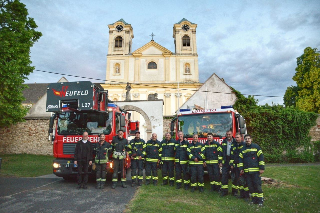 Eine Gruppe Feuerwehrleute posiert vor einer Kirche für ein Foto. Zwei Feuerwehrwagen sind vor ihnen geparkt.