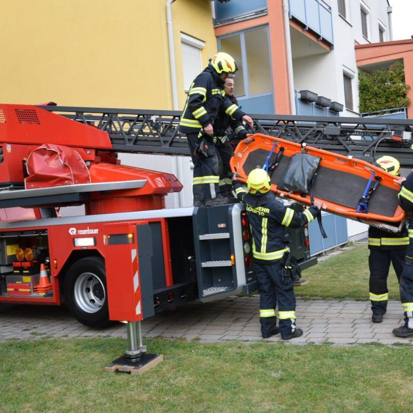 Feuerwehrleute stehen auf einer Leiter eines Feuerwehrwagens und halten eine Trage. Sie stehen vor einem Mehrfamilienhaus.