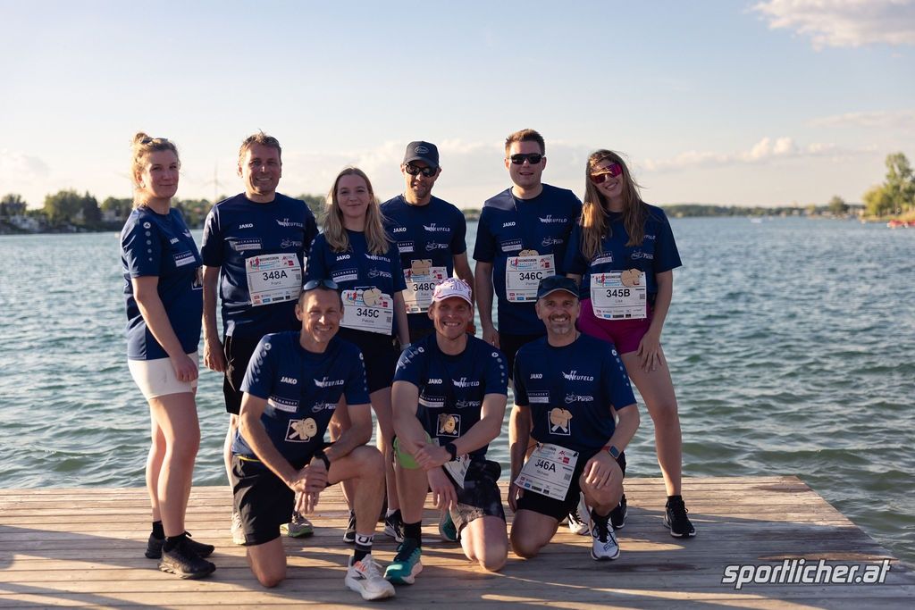 Eine Gruppe von Läufern posiert auf einem Steg am Wasser, alle tragen blaue Shirts und Startnummern. Sie lächeln für die Kamera.