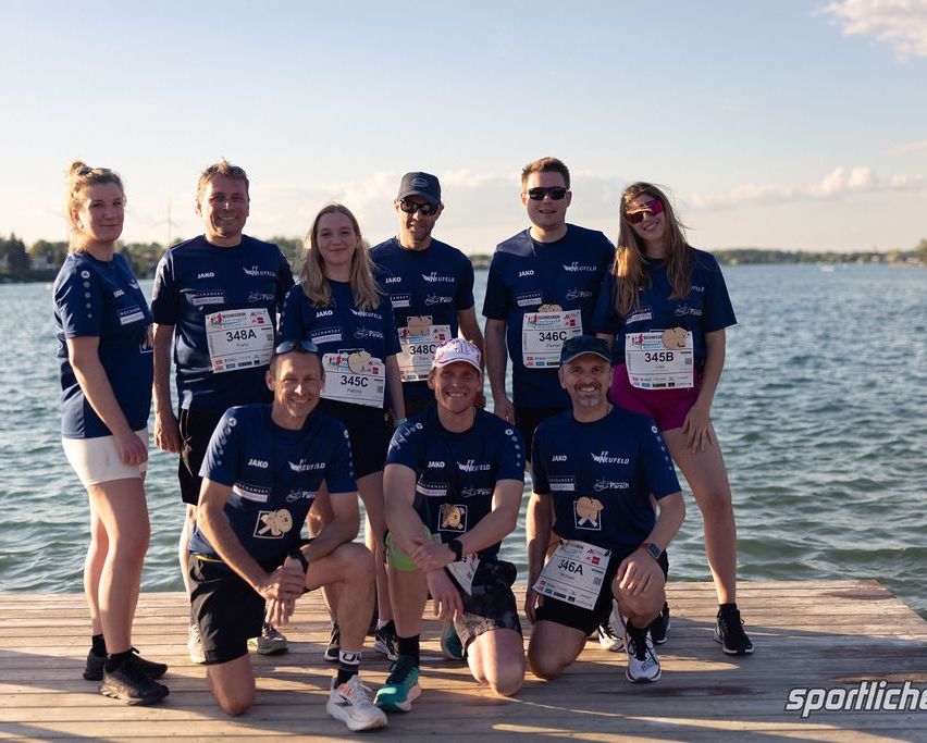 Eine Gruppe von Läufern posiert auf einem Steg am Wasser, alle tragen blaue Shirts und Startnummern. Sie lächeln für die Kamera.