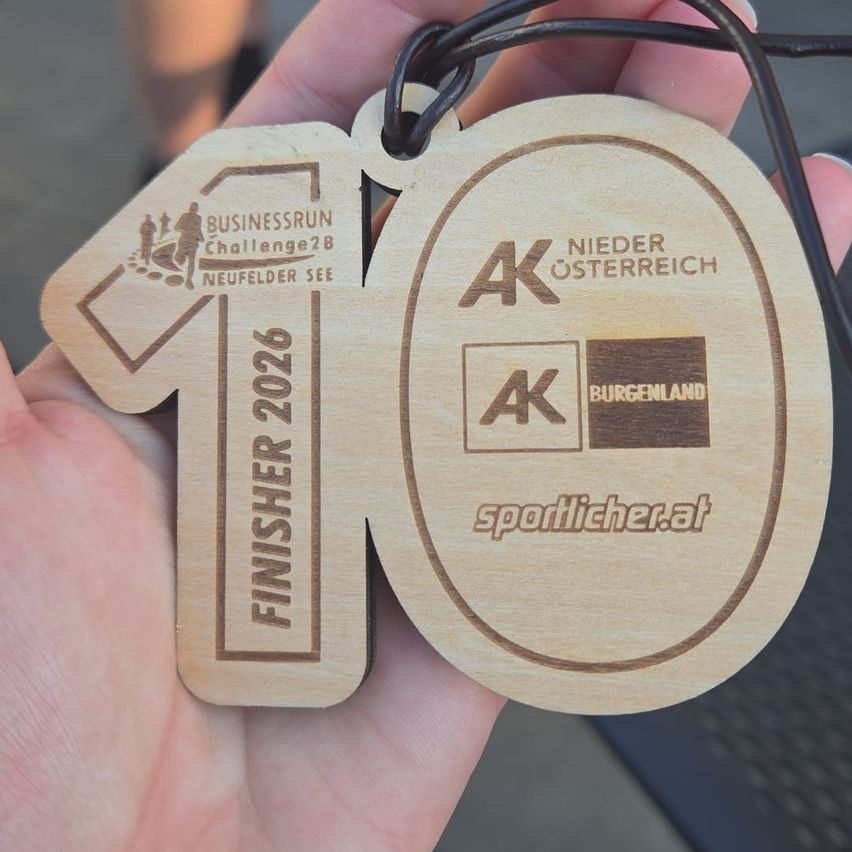 Eine hölzerne Medaille mit schwarzer Kordel, die ein Läufersilhouette und den Text 'FINISHER 2026' zeigt. Eine weitere Medaille zeigt 'NIEDER OSTERREICH' mit 'AK' und 'BURGENLAND' Logos. Die Website 'sportlicher.at' ist ebenfalls enthalten.