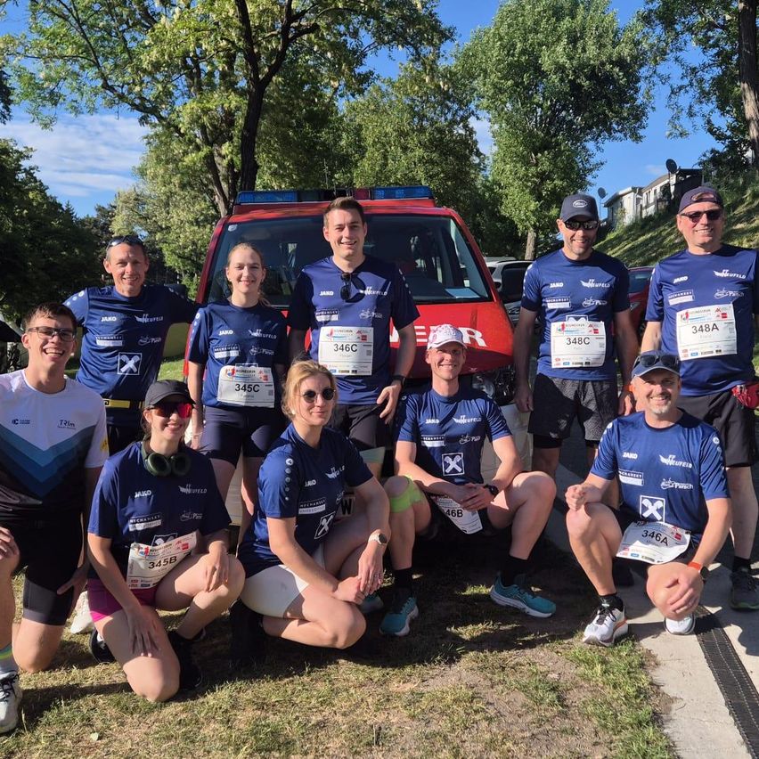 Eine Gruppe von Läufern in passenden blauen Trikots posiert für ein Foto mit einem roten Rettungswagen im Hintergrund. Bäume und ein klarer Himmel sind sichtbar.