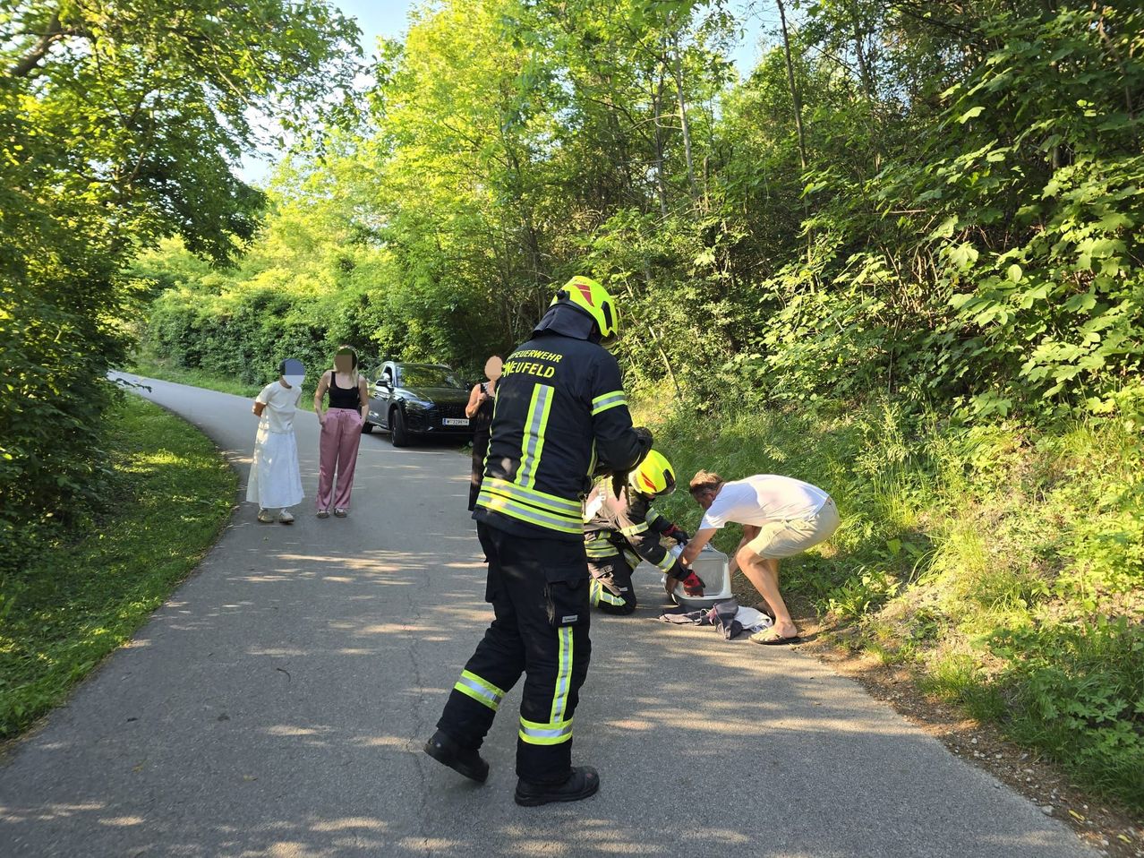 Feuerwehrleute helfen einem Mann auf einem Waldweg. Eine Frau in der Nähe hilft. Ein Auto ist am Straßenrand geparkt.