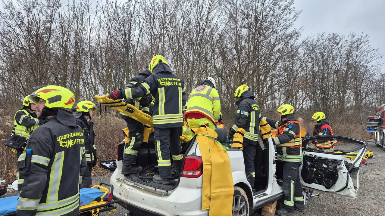 Eine Gruppe von Feuerwehrleuten in gelben und schwarzen Uniformen mit reflektierenden Streifen arbeitet an einem weißen Fahrzeug, möglicherweise einem Auto. Sie sind mit Helmen und Handschuhen ausgestattet.