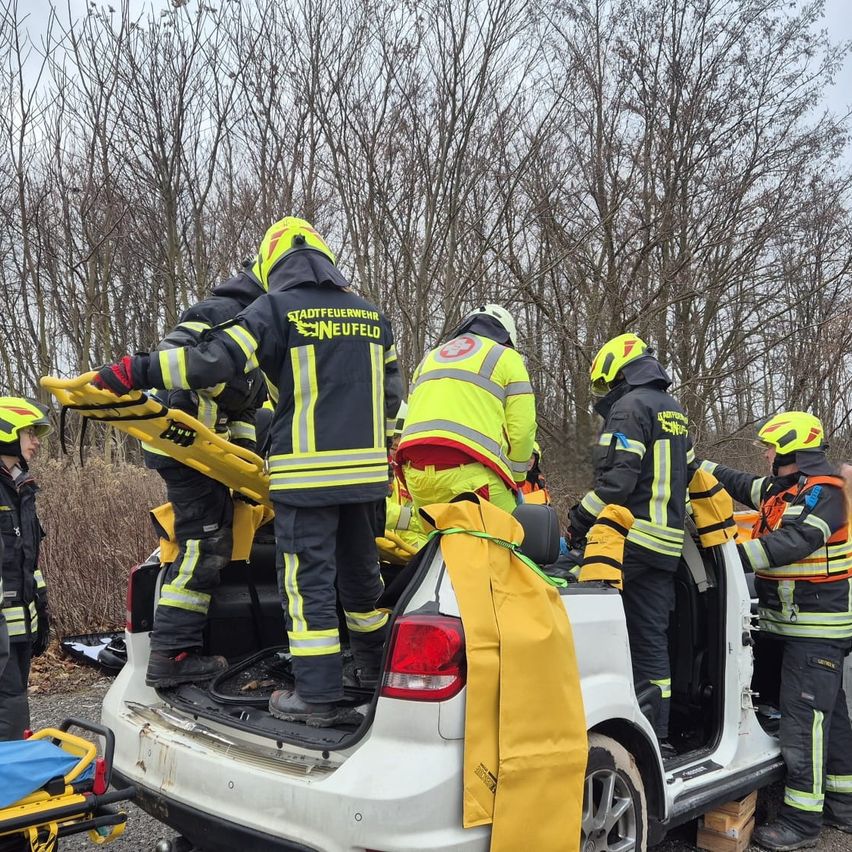 Eine Gruppe von Feuerwehrleuten in gelben und schwarzen Uniformen mit reflektierenden Streifen arbeitet an einem weißen Fahrzeug, möglicherweise einem Auto. Sie sind mit Helmen und Handschuhen ausgestattet.
