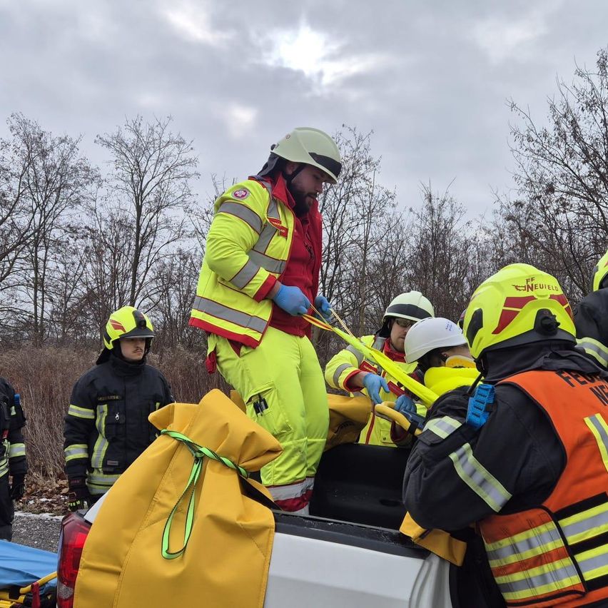 Rettungskräfte in gelben und roten Uniformen bereiten sich darauf vor, jemanden aus einem Fahrzeug zu extrahieren, wobei einer eine Trage hält und ein anderer auf dem Fahrzeugbett steht.