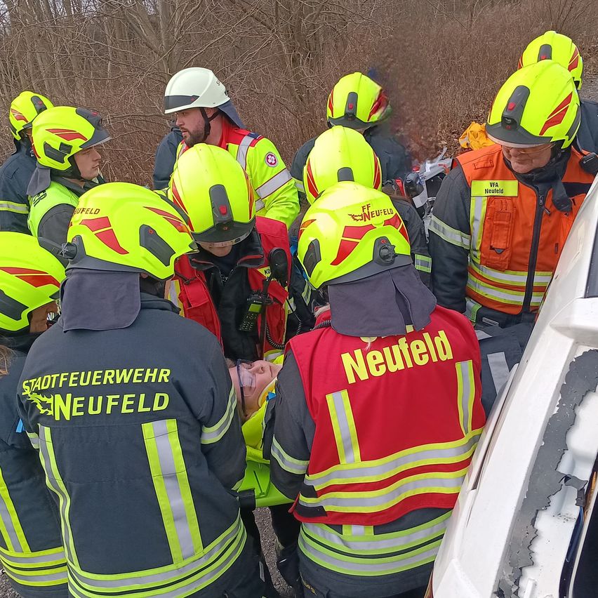 Rettungskräfte in gelben Helmen sind um eine Person in einer roten Weste mit der Aufschrift 'Neufeld' versammelt. Sie scheinen medizinische Hilfe zu leisten. Die Szene spielt sich im Freien ab, mit Bäumen im Hintergrund.