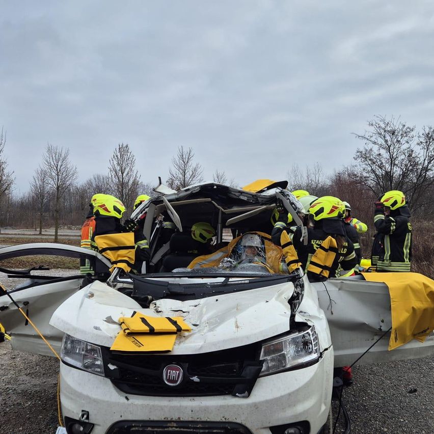 Rettungskräfte in gelben Helmen retten eine Person aus einem schwer beschädigten weißen Fiat SUV. Bäume und bewölkter Himmel im Hintergrund.