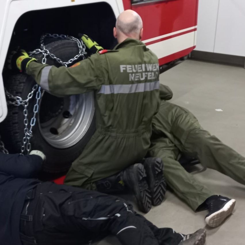 Ein Mann in einer grünen Uniform montiert Reifenketten an einem Fahrzeug, unterstützt von einer anderen Person. Das Fahrzeug steht in einer Garage geparkt.