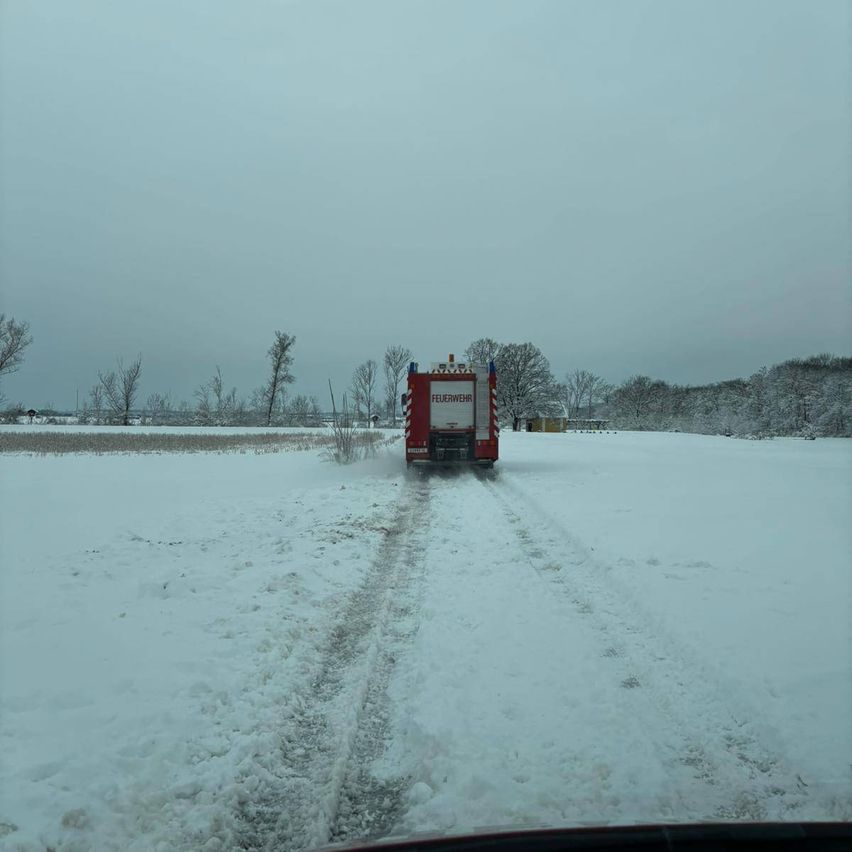 Ein roter Feuerwehrwagen fährt durch ein schneebedecktes Feld mit Bäumen in der Ferne. Der Himmel ist bedeckt.