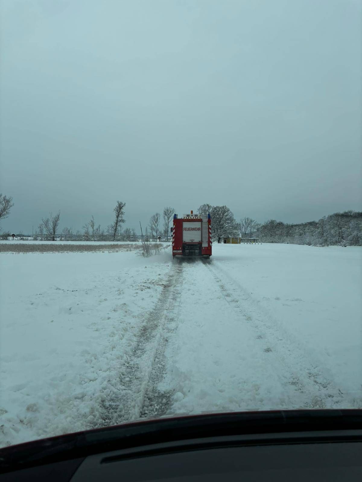 Ein roter Feuerwehrwagen fährt durch ein schneebedecktes Feld mit Bäumen in der Ferne. Der Himmel ist bedeckt.