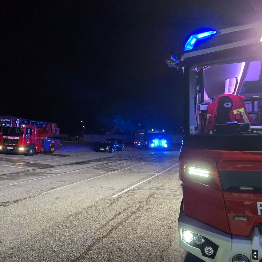 Feuerwehrfahrzeuge stehen nachts auf einer Straße, mit blinkenden Lichtern. Ein schwarzes Auto fährt in die entgegengesetzte Richtung.