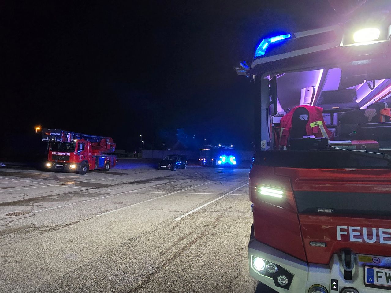 Feuerwehrfahrzeuge stehen nachts auf einer Straße, mit blinkenden Lichtern. Ein schwarzes Auto fährt in die entgegengesetzte Richtung.