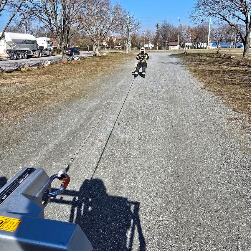Ein Feuerwehrmann zieht ein Seil, das an einem schweren Fahrzeug befestigt ist, auf einer Schotterstraße, in der Lkws und Autos parken.