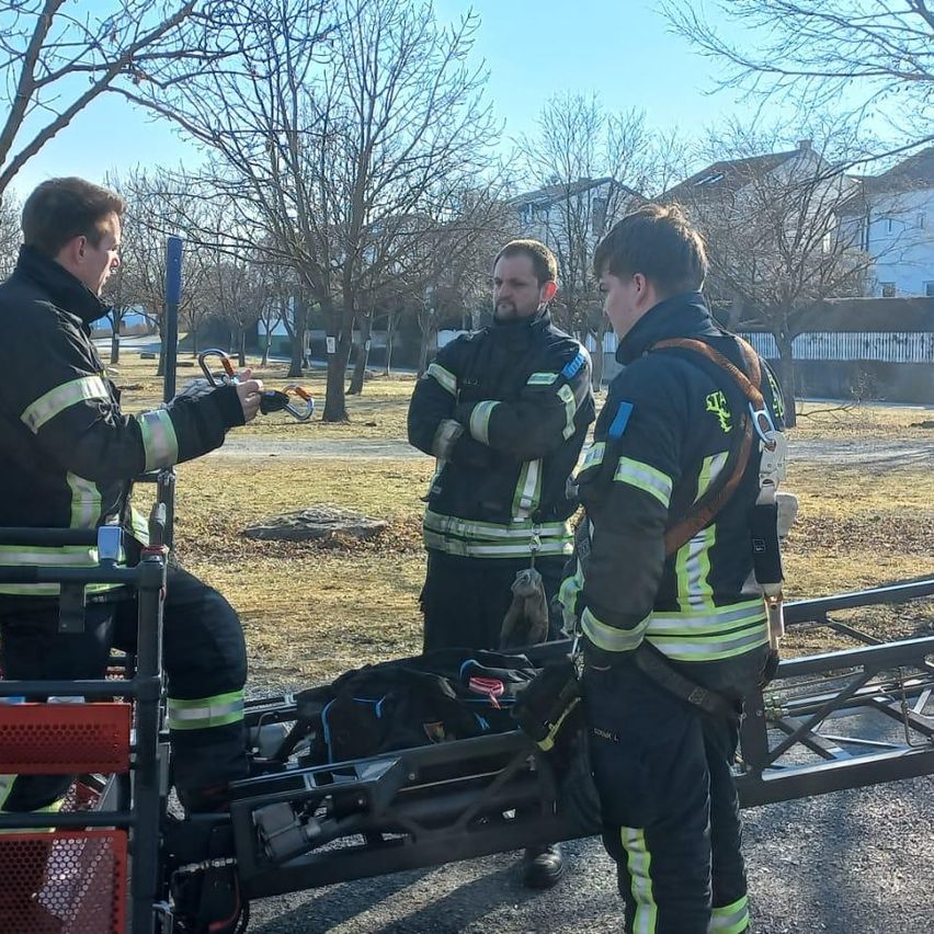 Drei Feuerwehrleute in Uniform stehen in einem Außenbereich in der Nähe eines Feuerwehrwagens, einer hält ein Werkzeug. Sie stehen in der Nähe eines Trolleys mit Taschen darauf. Im Hintergrund befinden sich trockene Bäume und Gebäude.