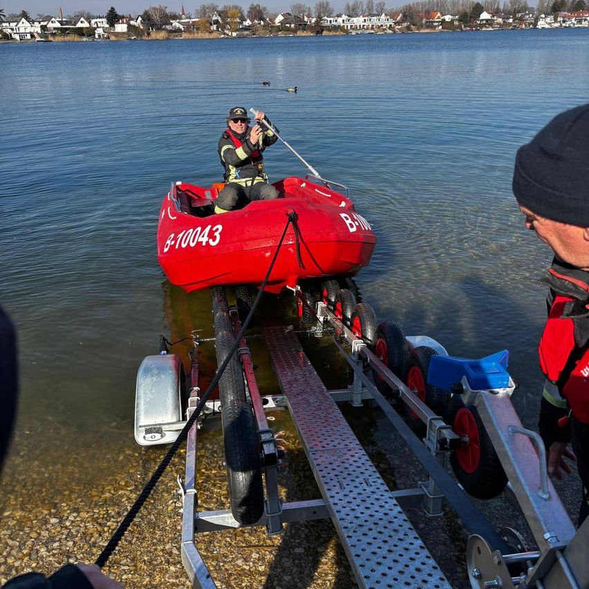 Eine Person in einem roten Boot auf einem Anhänger, bereit für eine Wasserrettung. Eine weitere Person steht daneben, und Häuser und Bäume sind im Hintergrund zu sehen.