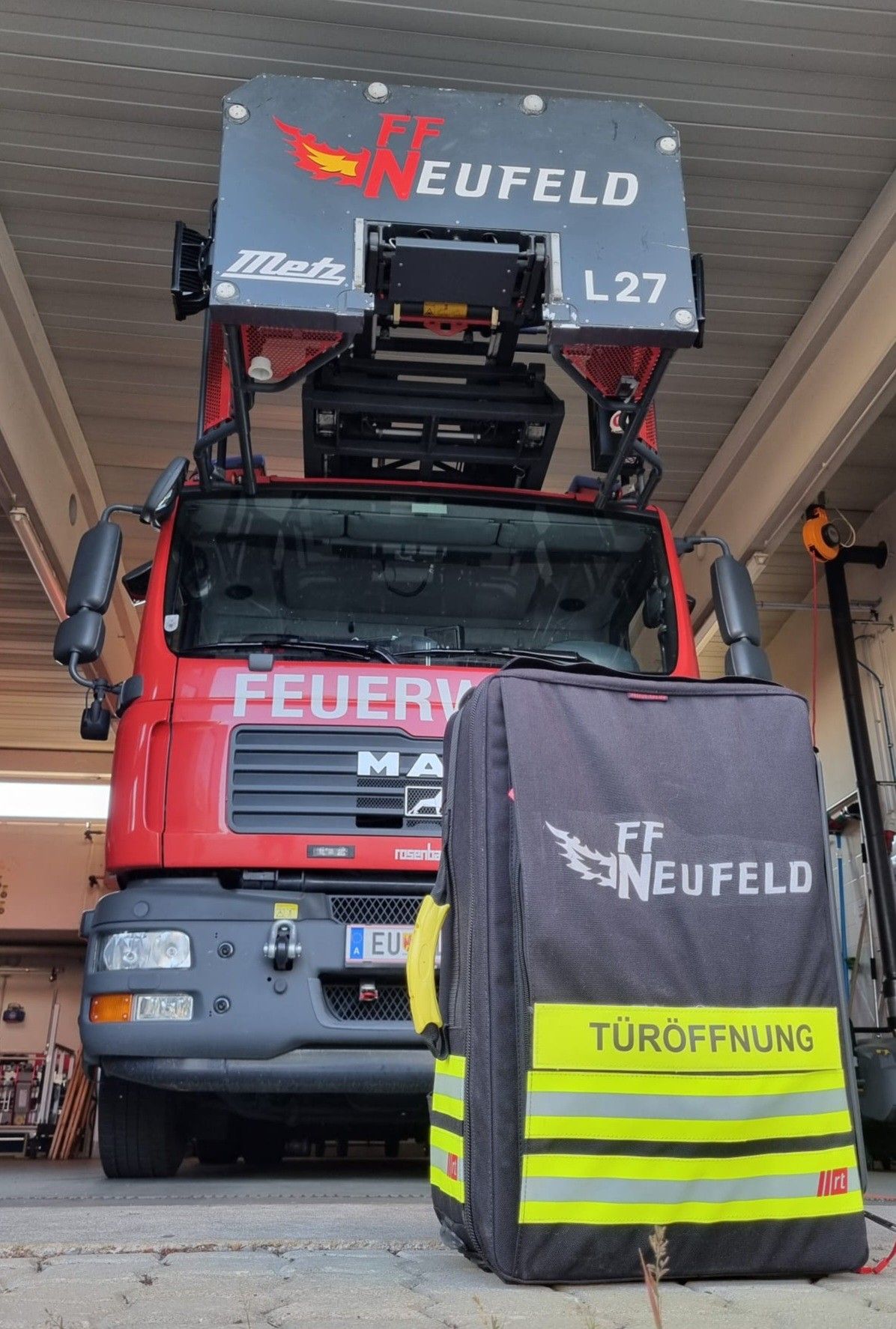 Ein roter Feuerwehrwagen mit 'FEUERW' auf der Front, ein Rucksack mit 'FF NEUFELD' und 'TÜRÖFFNUNG' darauf. Der Wagen hat einen großen Kran oben mit 'Metz L27'.
