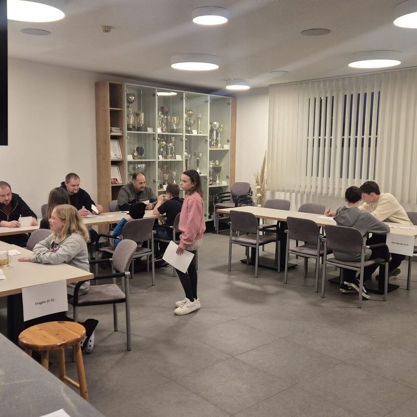 Eine Gruppe von Menschen sitzt an Tischen und schreibt. Ein Mädchen steht und hält Papiere. Trophäendisplays sind hinter ihnen. Der Raum hat geflieste Böden und Deckenleuchten.