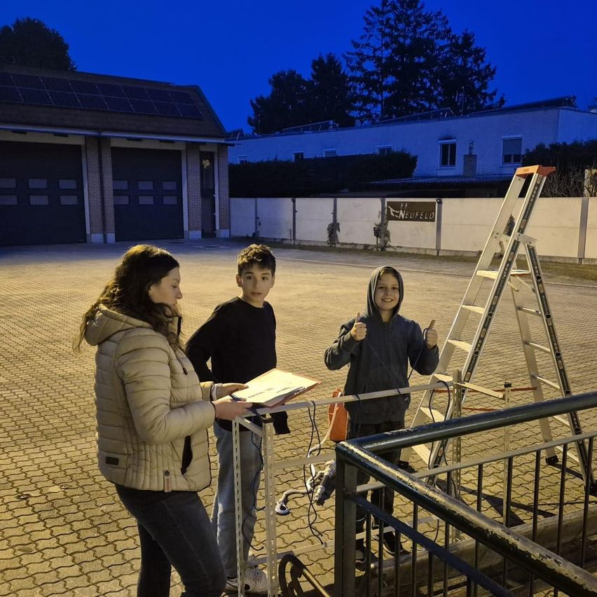 Drei Personen stehen im Freien. Eine Frau und zwei Jungen posieren für ein Foto. Die Frau hält ein Buch und steht in der Nähe eines Zauns. Dahinter befindet sich ein Gebäude mit einer Garage und einer Leiter.
