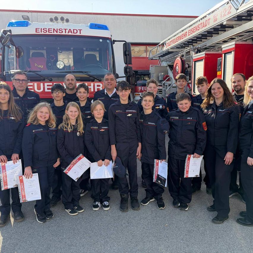 Eine Gruppe junger Feuerwehrleute in Uniform posiert vor einem mit Eisenstadt beschrifteten Feuerwehrwagen. Sie halten Urkunden in den Händen und lächeln, während Erwachsene hinter ihnen stehen.