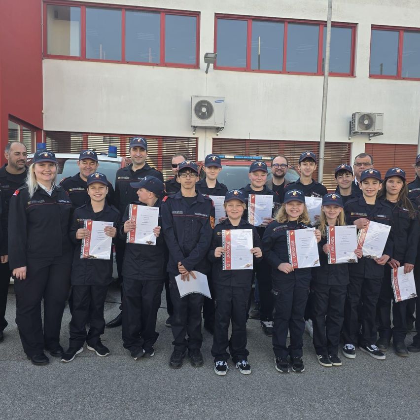 Eine Gruppe junger Personen in Uniform, die Urkunden halten, posiert für ein Foto vor einem Gebäude mit Glasfenstern und rotem Rand.