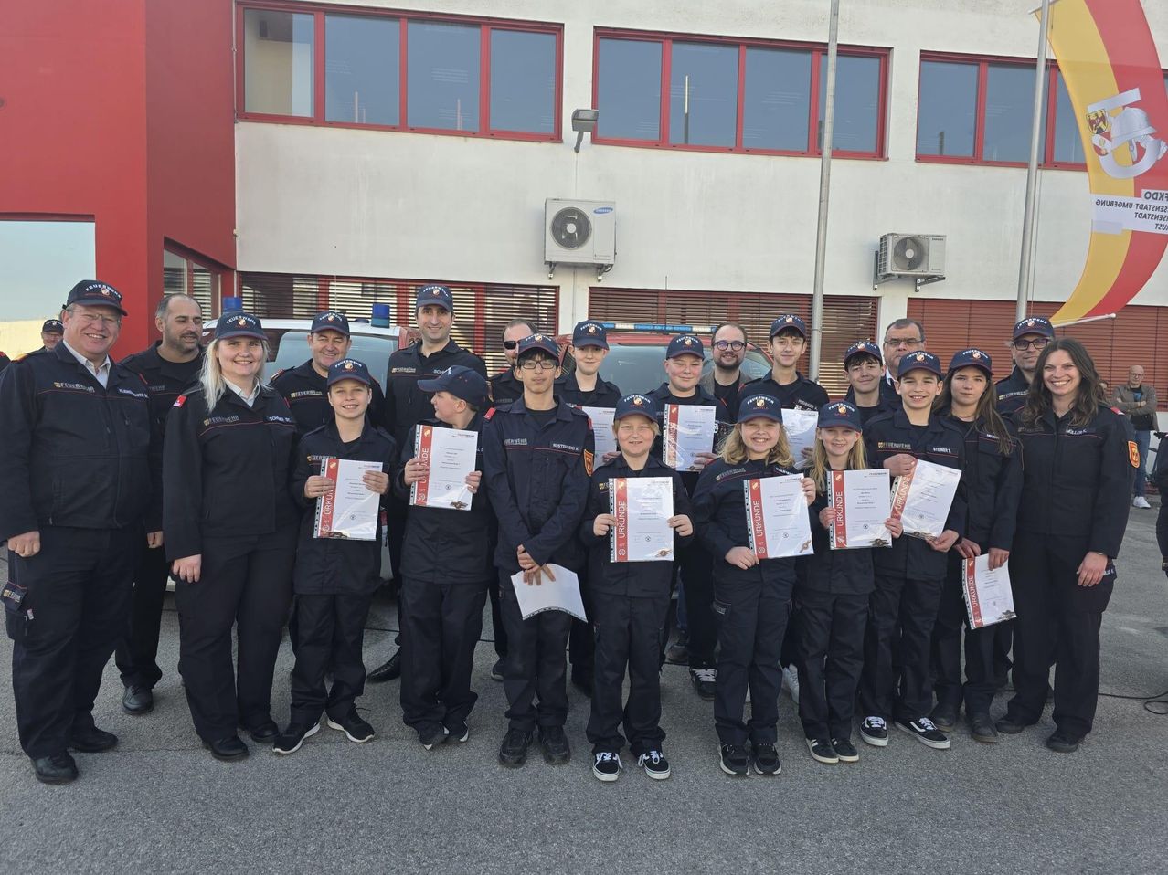 Eine Gruppe junger Personen in Uniform, die Urkunden halten, posiert für ein Foto vor einem Gebäude mit Glasfenstern und rotem Rand.