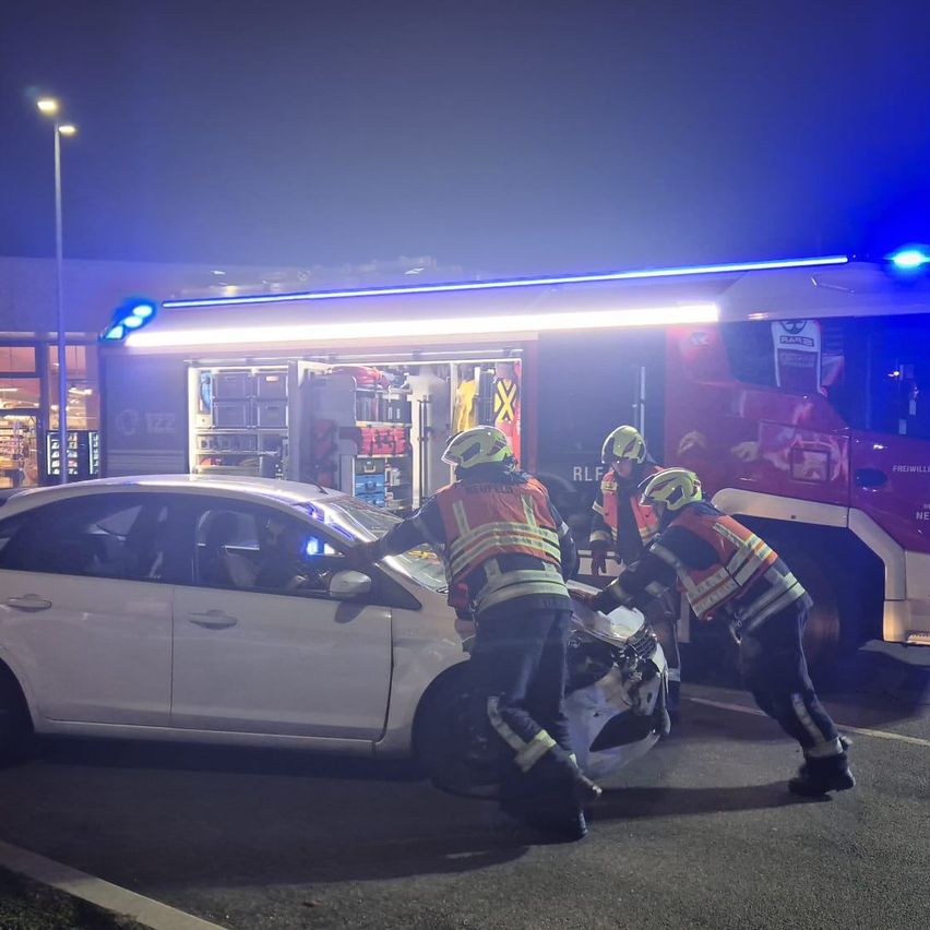 Drei Feuerwehrleute in Warnwesten helfen einer Person nach einem Autounfall bei Nacht, mit einem Feuerwehrwagen und einem weißen Auto im Hintergrund.