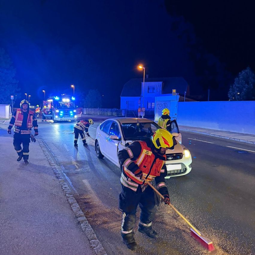 Einsatzkräfte reinigen bei Nacht eine Verschüttung auf der Straße. Sie tragen Schutzausrüstung und benutzen Besen. Ein Auto ist in der Nähe geparkt und ein Feuerwehrwagen ist im Hintergrund.