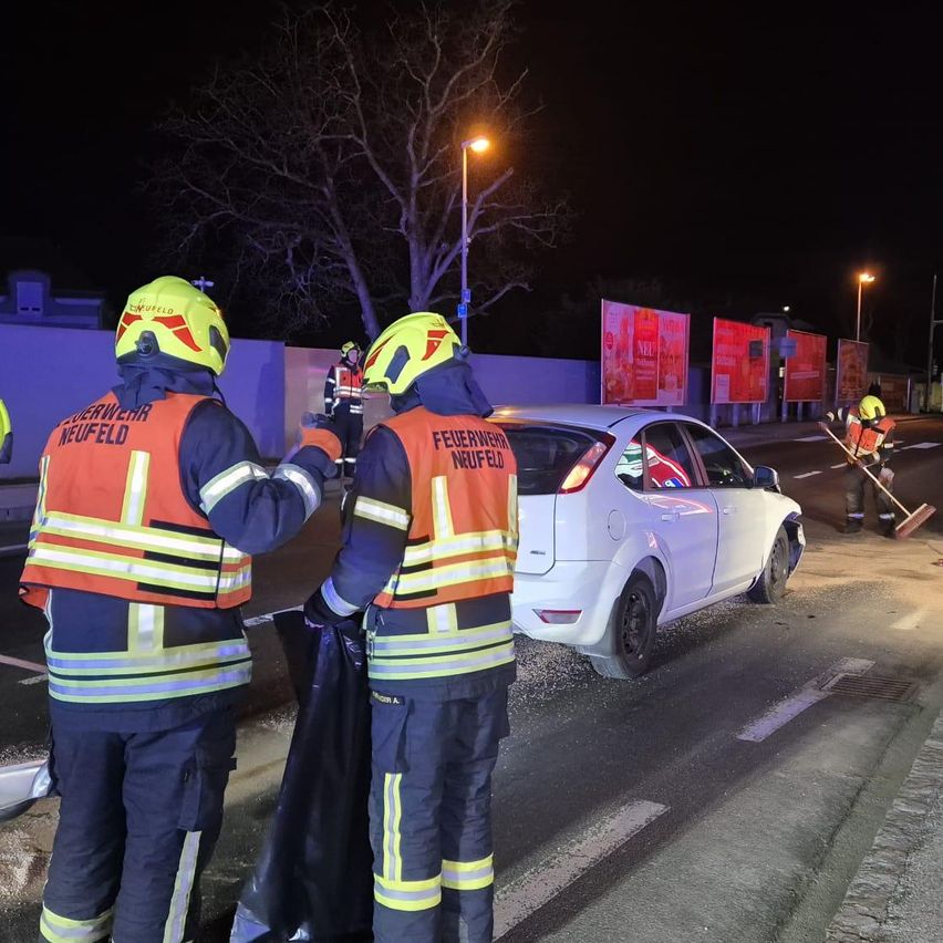 In der Nacht putzen zwei Feuerwehrleute in orangefarbenen Westen mit 'Feuerwehr Neufeld' die Straße. Ein weißer Wagen ist am Straßenrand geparkt, und ein dritter Feuerwehrmann fegt die Straße.