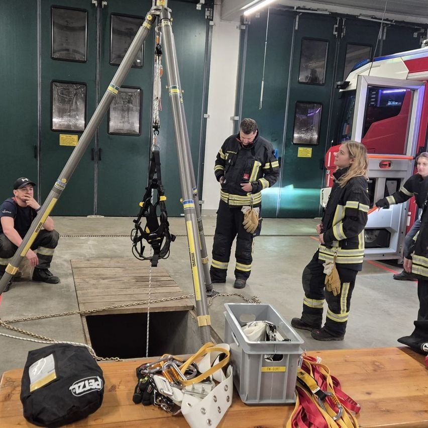 Feuerwehrleute üben Rettungsaktionen in einer Garage. Ein Feuerwehrmann kniet neben einer Falltür, ein anderer steht bereit mit einem Seil und Gurtzeug. In der Nähe ist ein Feuerwehrauto geparkt.