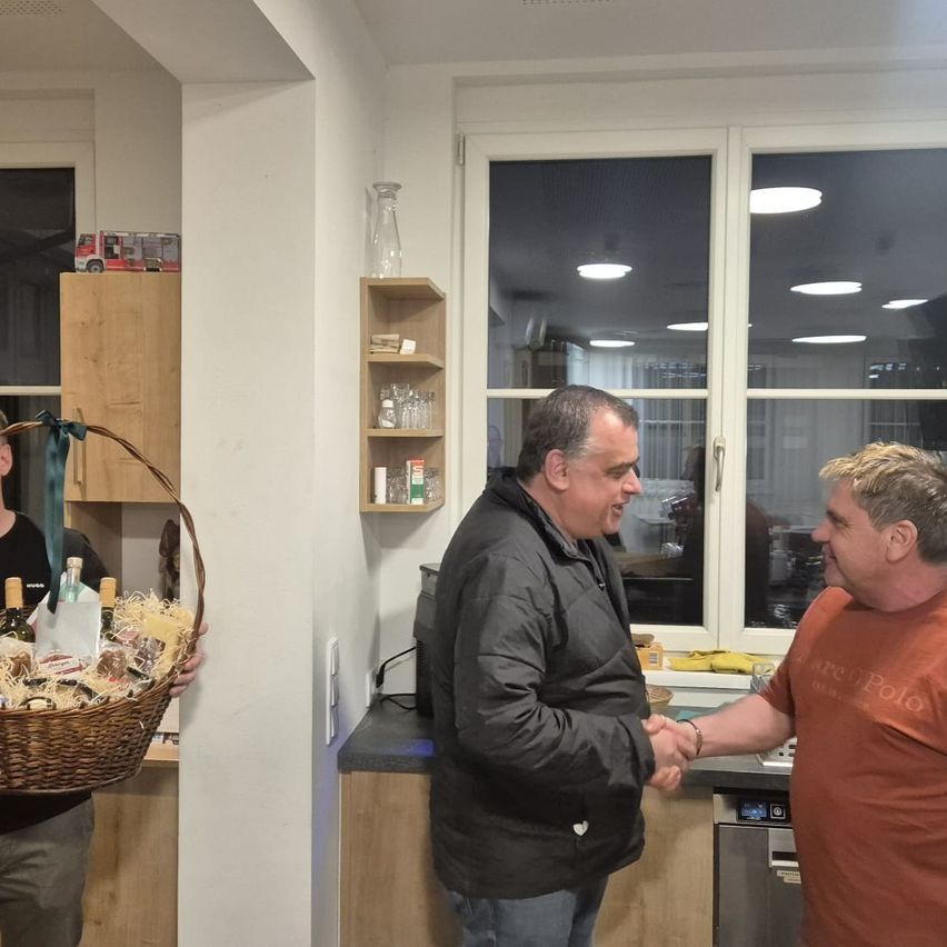 Zwei Männer schütteln sich in einer Küche die Hände. Ein Mann hält einen Korb mit verschiedenen Gegenständen. Ein anderer Mann trägt eine Jacke. An der Wand steht ein Regal mit Gegenständen.