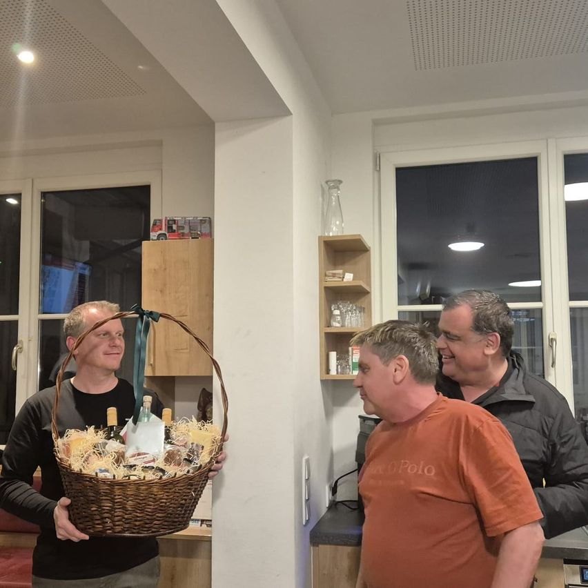 Drei Männer stehen in einer Küche, einer hält einen Korb mit verschiedenen Lebensmitteln. Ein anderer Mann schaut in den Korb, während der dritte Mann lächelt. Im Hintergrund befinden sich Schränke und Regale.