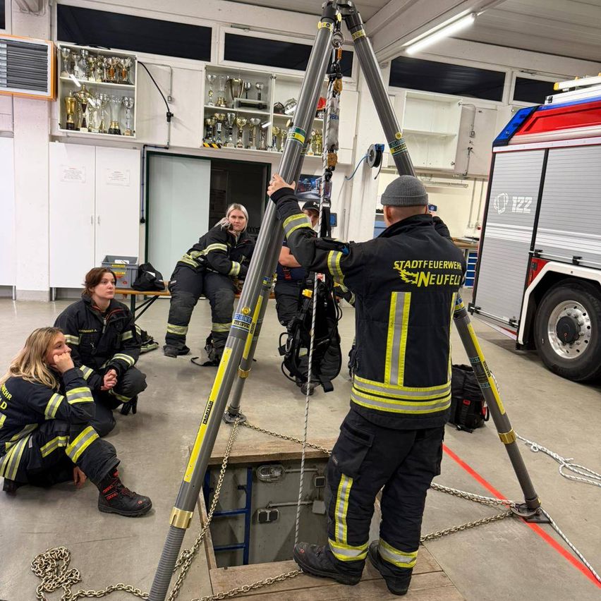 Eine Gruppe von Feuerwehrleuten trainiert in einem Raum, in dem ein Feuerwehrwagen geparkt ist. Eine Person steht auf einer Plattform, während andere sitzen und zusehen.