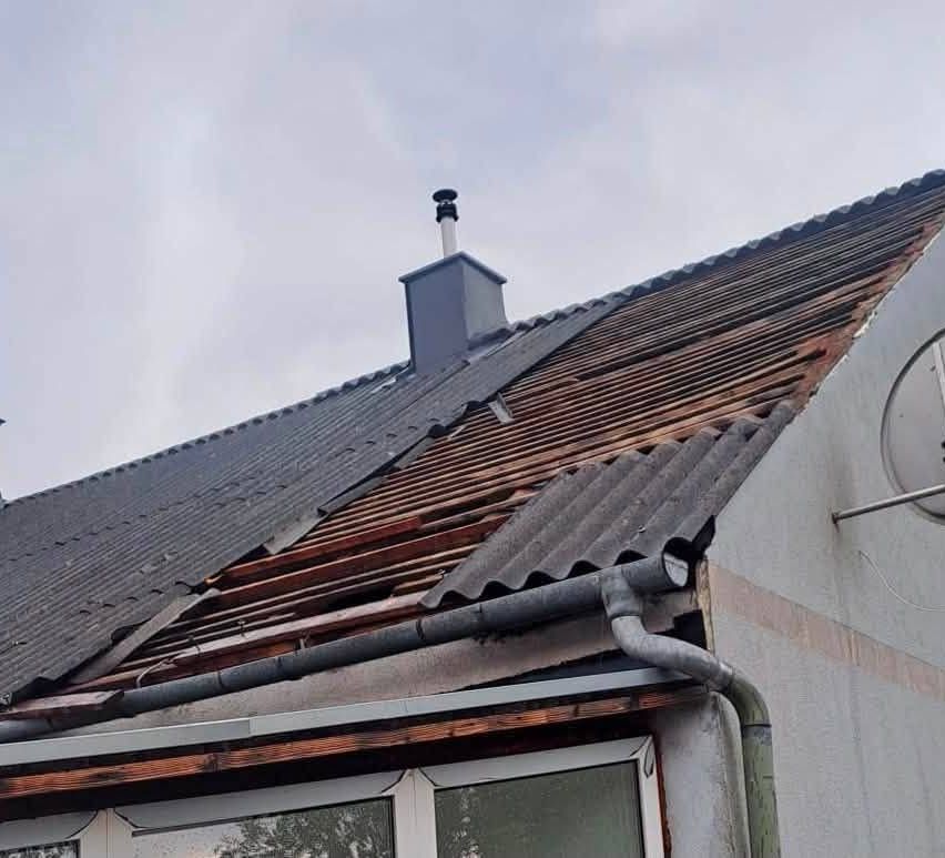 Ein Haus mit einem beschädigten Dach und einem Schornstein darauf. Das Dach besteht aus Metallblechen und hat einen Dachrinnen an der Seite. Das Haus hat ein Glasfenster und eine Tür.