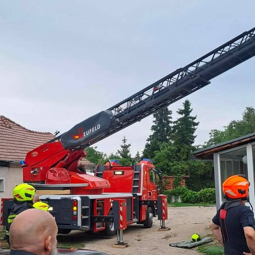 Ein roter Feuerwehrwagen mit dem Wort Eufeld auf seiner Leiter steht vor einem Haus. Zwei Feuerwehrleute stehen in der Nähe des Trucks und ein Mann steht vor dem Haus.