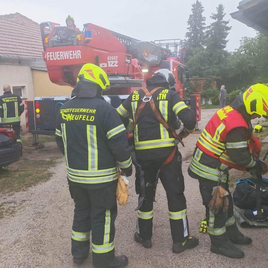 Drei Feuerwehrleute in gelben und schwarzen Uniformen stehen auf einem Kiesweg, einer justiert seinen Gurt. Ein Feuerwehrwagen ist hinter ihnen geparkt.