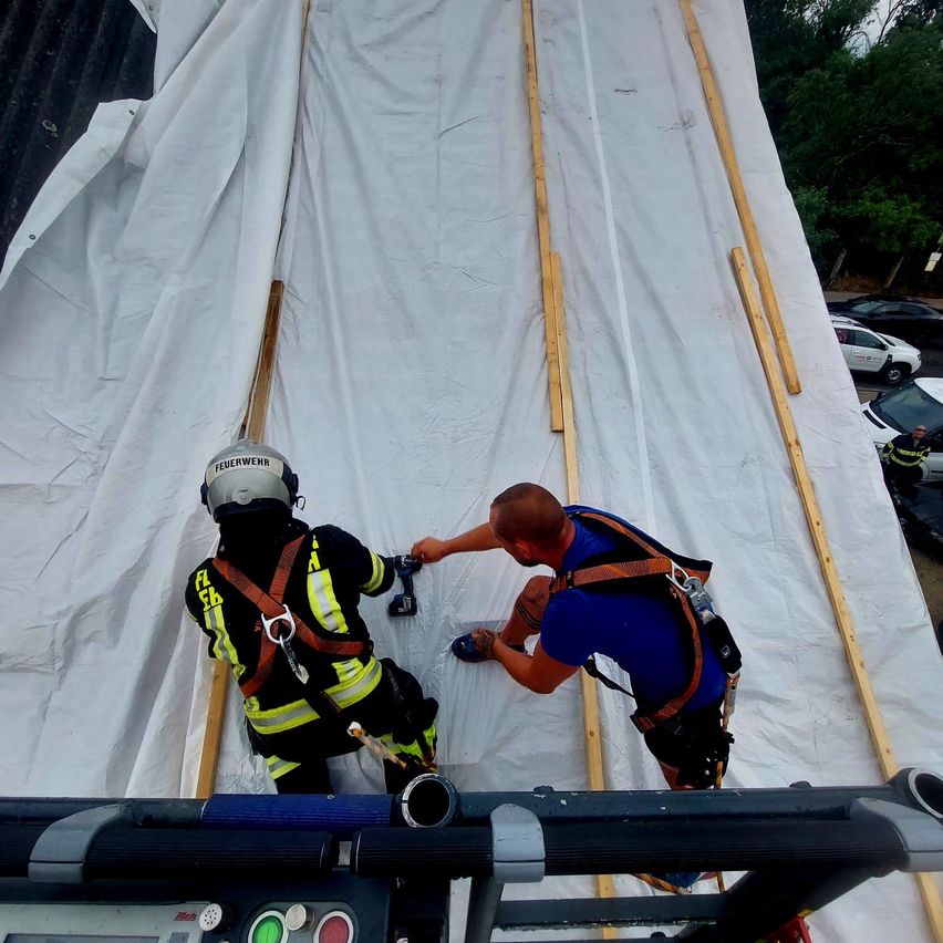 Zwei Feuerwehrleute arbeiten an einem großen weißen Planen, einer hält ein Werkzeug, während der andere hilft. Sie tragen beide Schutzausrüstung und stehen auf einer Plattform.