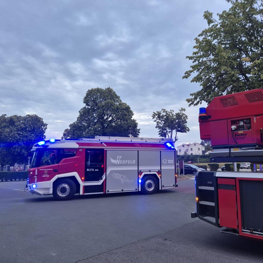 Ein roter Feuerwehrwagen mit dem Wort Neufeld an der Seite fährt auf einer Straße mit Bäumen und einem bewölkten Himmel im Hintergrund.
