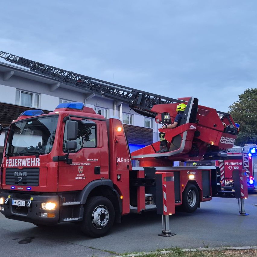 Ein roter Feuerwehrwagen mit ausgefahrener Leiter steht vor einem Gebäude. Der Fahrer ist in der Kabine, und ein Feuerwehrmann steht auf der Leiter.