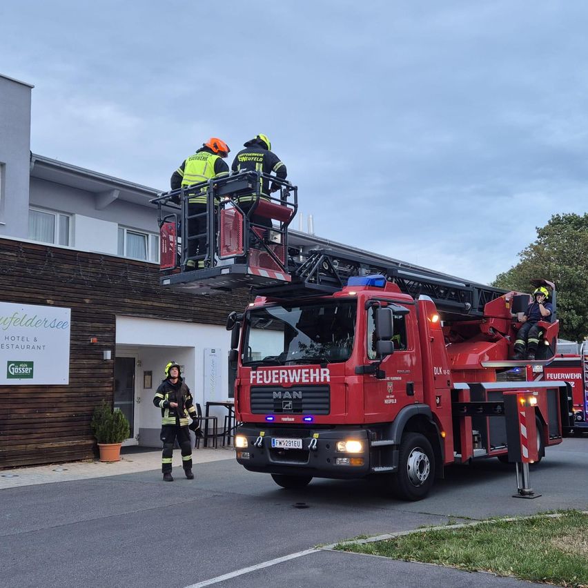 Feuerwehrleute stehen auf einer Leiter, die an einem roten Feuerwehrauto befestigt ist, das vor einem Gebäude mit dem Schild 'Hotel & Restaurant' parkt. Ein Feuerwehrmann sitzt auf der Leiter, während ein anderer am Boden steht.