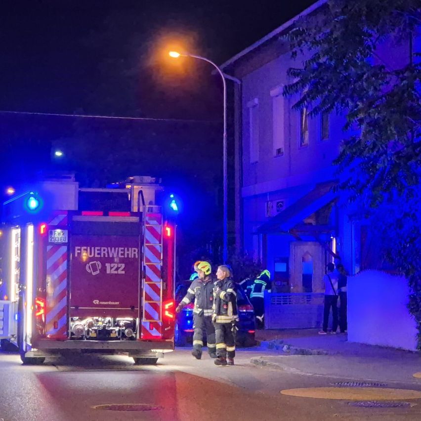 Zwei Feuerwehrleute in voller Ausrüstung stehen vor einem Feuerwehrwagen mit blinkenden Lichtern, in der Nähe eines Gebäudes mit beleuchtetem Eingang und einer Straßenlaterne.