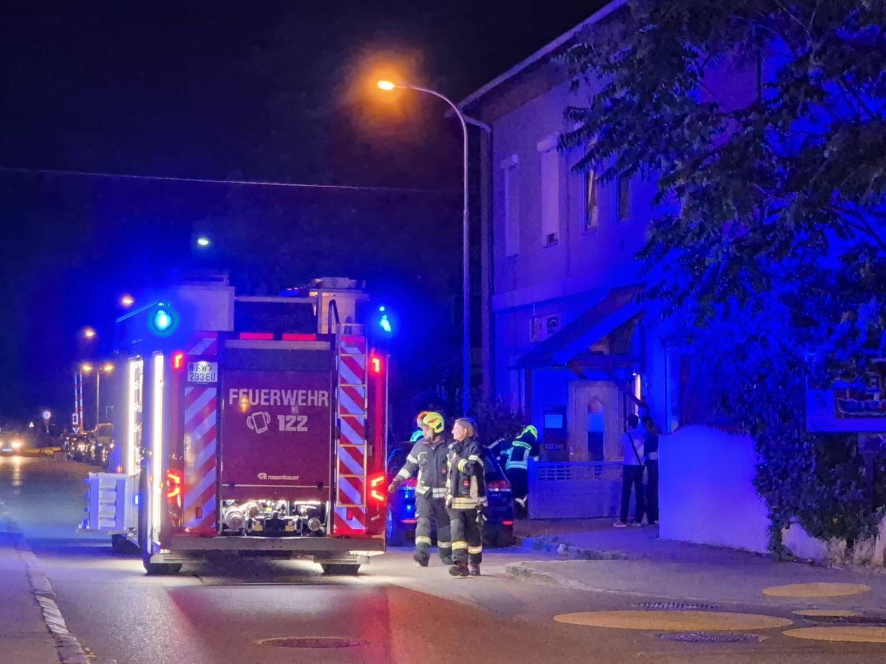 Zwei Feuerwehrleute in voller Ausrüstung stehen vor einem Feuerwehrwagen mit blinkenden Lichtern, in der Nähe eines Gebäudes mit beleuchtetem Eingang und einer Straßenlaterne.