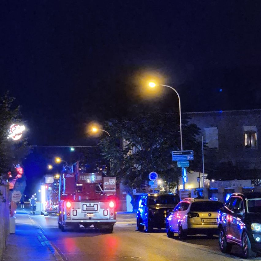 Ein Feuerwehrauto mit blinkenden Lichtern steht nachts auf der Straße, mit mehreren anderen Fahrzeugen in der Nähe. Straßenlaternen und Schilder sind im Hintergrund zu sehen.