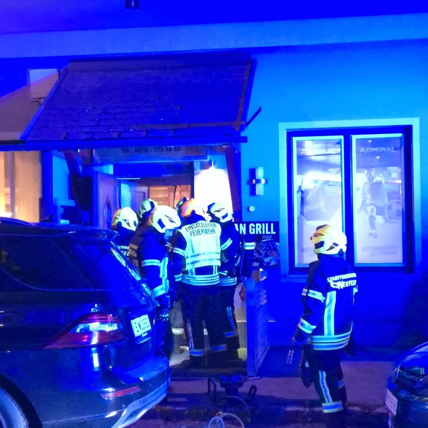 Feuerwehrleute in Schutzkleidung stehen vor einem blau beleuchteten Gebäude, möglicherweise als Reaktion auf einen Notfall. Ein beschädigtes Auto ist in der Nähe geparkt.