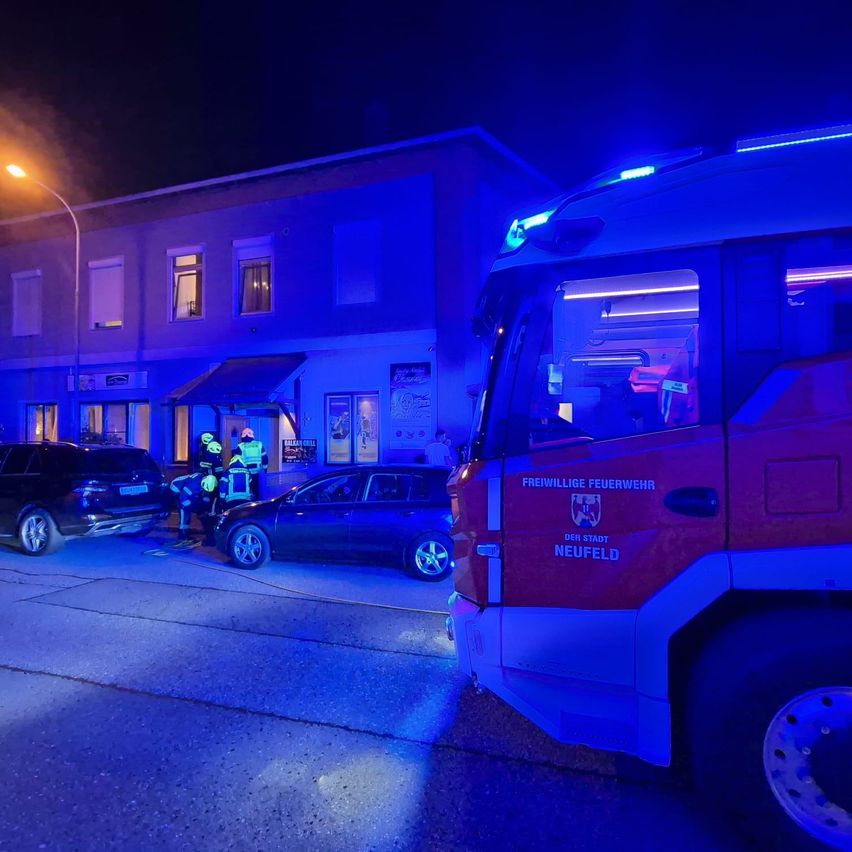 Ein roter Feuerwehrwagen mit blinkenden blauen Lichtern steht nachts auf der Straße. Zwei Personen in gelben Helmen stehen neben einem beschädigten schwarzen Auto. Ein Gebäude mit blauen Lichtern ist im Hintergrund.